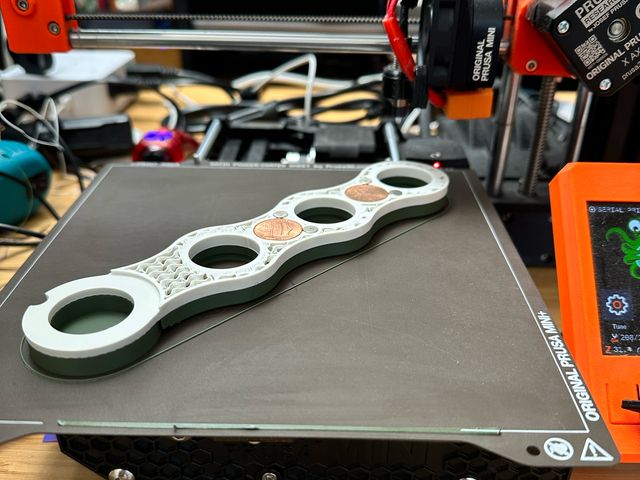 A 3D printer with a print in progress on the bed. The print is wavy and green on the bottom, but the upper layers that are printing now are white. The inside is filled with a wavy pattern of plastic, known as "gyroid infill." There are holes for the dowel mounts to snap into but between those holes are holes in which pennies and cylindrical nickel plated magnets have been embedded.