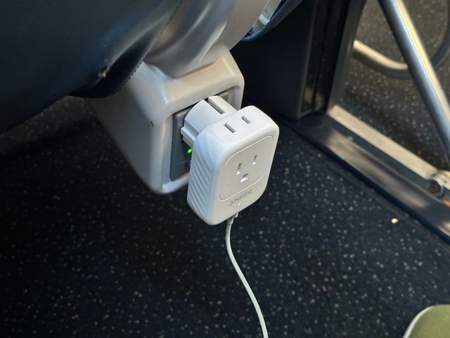 A larger white Anker brand adapter plugged into an under-seat airplane power outlet. On its face a three prong U.S.-style outlet is available. On its top is a two prong outlet. From beneath it a thin white USB charging cable extends.