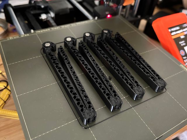 A batch of 4 clips on a 3D printer bed, paused mid-print. Four shiny circular magnets are slotted into holes at the head of each clip