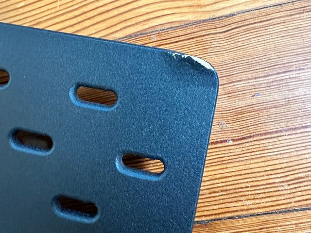 An IKEA Skådis pegboard with the corner scuffed and the brown of the particleboard showing through a tear in the black finish.
