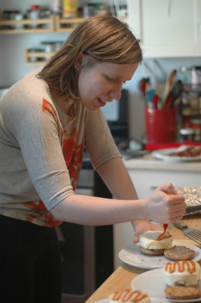 A blond woman with a focused smile adding a zig zag drizzle of hot sauce to the white melted cheese of a breakfast sandwich.