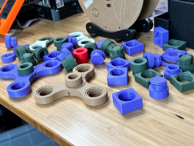 A chaotic pile of 3D printed parts of different colors all on a table. It's a mix of cylindrical parts with knurled caps and different connectors—screws, snaps, and twist lock—and rectangular blocks with holes in the middle as well as a couple complete mini whistle stands with only four holes. Some parts appear to have failed midway through and are either missing bits or degrade into stringy plastic on one end.