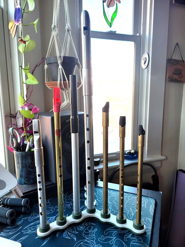 The whistle stand now on a desk with a blue desk mat and plants in the background. There are tin whistles over each of the dowels standing straight up with the tallest in the middle and the shortest on the outsides.
