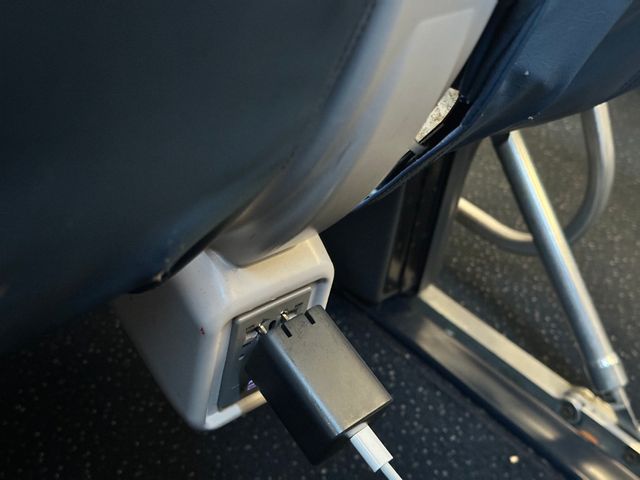 A small cubical dark gray USB adapter brick plugged into an under-seat airplane power outlet. It is loose and has fallen out slightly, exposing the silver metal of its two prongs.