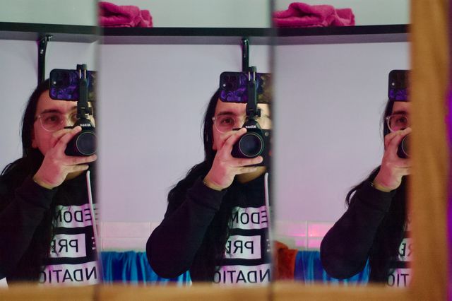A photo of myself reflected three times in a divided bathroom mirror. The light has a purple tint. I am holding a Nikon DSLR with an iPhone mounted to the top with a black plastic phone mount.