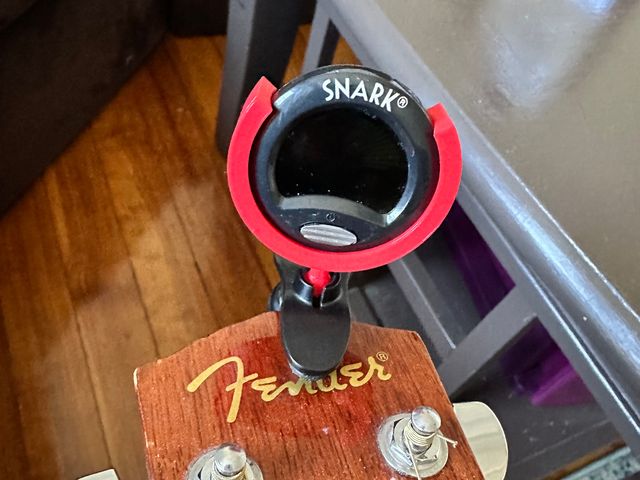 A Snark guitar tuner clipped onto a guitar headstock. A red plastic semicircle surrounds the circumference of the tuner's round display and from it extends a red ball joint secured in the clip.