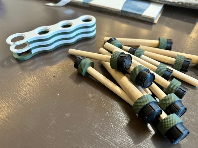 The whistle stand disassembled on a table. It is made up of three wavy legs stacked on top of each other and in front of them a pile of wooden dowels mounted on green cylinders with black tabbed snap connectors at the bottom