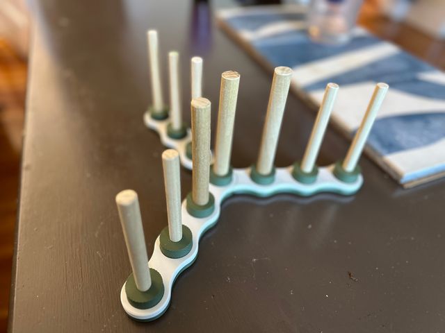 A table with a large green and white plastic object on it. The object has three wavy legs and evenly spaced along each leg is a green cylindrical mount with a short wooden dowel protruding from the top. It looks a bit like a multidimensional menorah. On the table are three spare dowels with green topped mounts attached to the end.