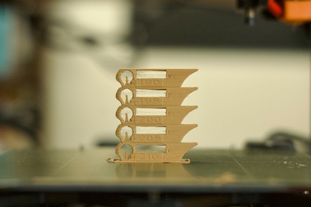 A layered tan object sitting on a green 3D print bed. The object is a shape with a variety of features designed to test different 3D printing qualities. It is repeated vertically 5 times, labeled with a temperature on each layer, ranging from 210 at the bottom to 190 at the top.