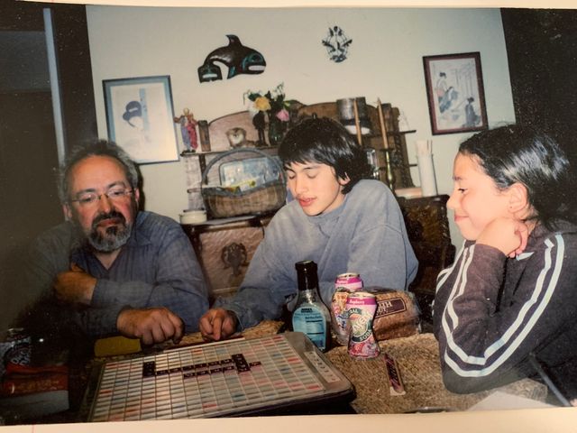 Three people around a Scrabble board on a dining table. On the left, an older man with glasses and a gray-streaked beard. In the center, a teenage boy in a gray sweatshirt looks down at the tiles. On the right, a younger girl smiles slightly.