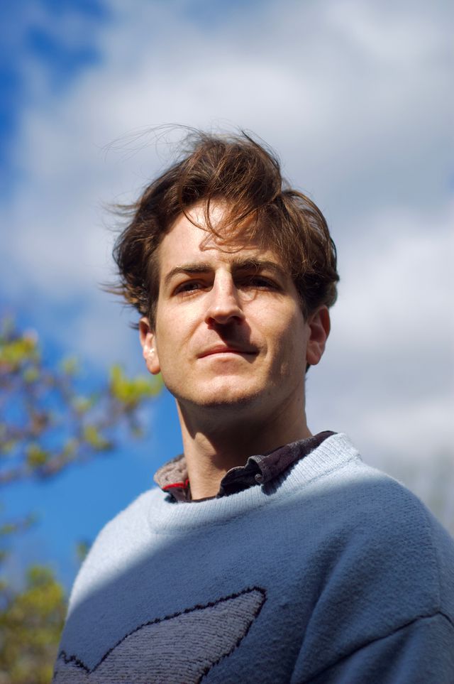 Close up portrait of a 30-something white man taken from slightly below. Behind him the sky is blue. A shadow crosses his blue knit sweater dramatically.