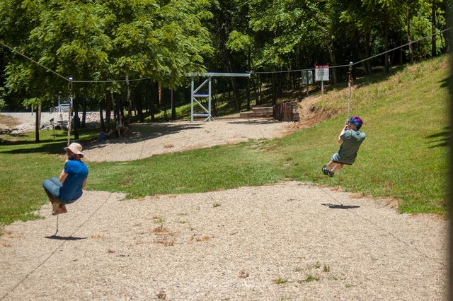 Two adults on parallel ziplines in a green park with gravel beneath them. It's a sunny day.