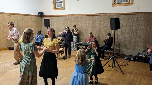 A room with contra dancers holding two hands with their partner, in motion. In the background I'm calling with a band.
