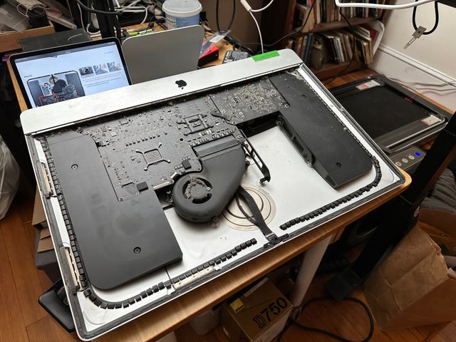 An iMac chassis resting on its back on a table. The glass screen has been removed, exposing a number of black circuitboards and black plastic parts. The internals are a bit dusty.