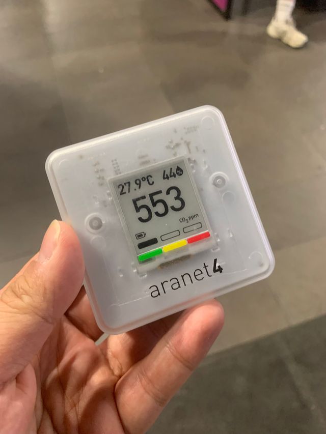 Photo of a hand holding an aranet4 device. The device is small, square, made of translucent white plastic that reveals the inner electronics. It has an e-ink screen that shows the temperature, humidity, and CO₂ concentration in PPM.
