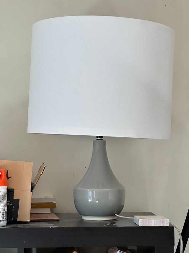 A gray ceramic lamp on a shelf with a sligthly lopsided oversized lampshade over the top.