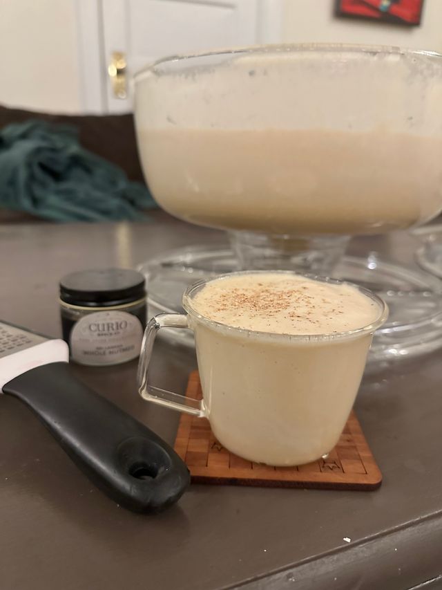 A glass mug full of eggnog, thick and creamy with nutmeg on top. Beside it is a spice jar of whole nutmeg and a grater. In the background, out of focus, is a punch bowl full of creamy, foamy, nog.