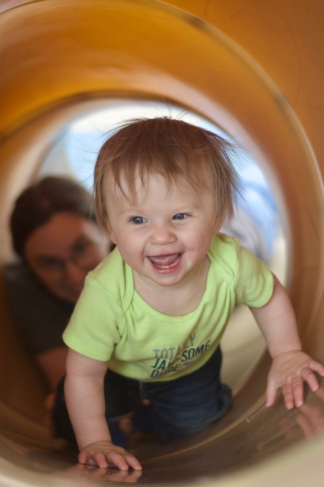 A small blonde child is crawling through a light brown play structure tunnel toward the camera. She's grinning and excited. Behind her, out of focus, her mother is reaching toward her and smiling.