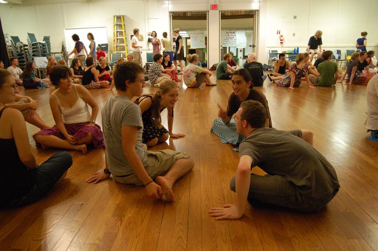 Dozens of young people sitting in small groups on the wood floor of a large hall, talking. In the foreground a dancer in a brown shirt and teal skirt leans in, gesturing, toward a person in glasses kneeling across from them.