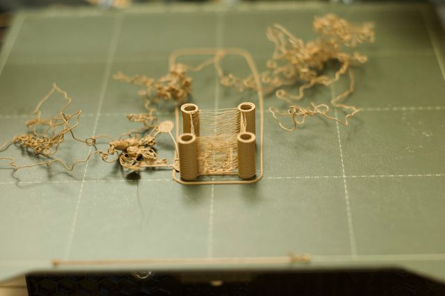 Four tan threaded cylinders sitting on a green 3D print bed. There's stringy plastic between the cylinders all the way up the vertical height. Int the background is a loose accumulation of extruded plastic in a curly clumped strand.