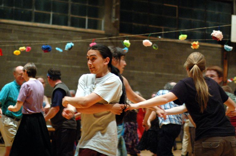 A young dancer with long dark hair in a white t-shirt, circling with other dancers in a gym hung with strings of paper flowers. Their hands are joined with their neighbors' but their arms are awkwardly crossed in front of their body, and they're wide-eyed and grimacing over their shoulder.