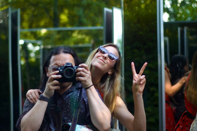 A photo shot amidst an array of mirrors. Green foliage is visible in the background. In the mirror directly facing the camera is the photographer, an Asian man, with long dark hair and a black shirt, deep in focus, face behind his Nikon camera. Behind him, with her arm around him, is a white woman with long blonde hair and a red dress in sunglasses and throwing up a peace sign.