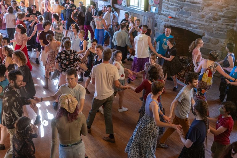 A crowded wooden dance floor seen from above, with dozens of young contra dancers engaged mid-dance, taking hands in rings of four. A big stone fireplace fills one wall, and sunlight from the windows gleams on the floor.