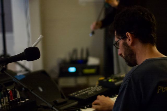 A bearded man at a keyboard, mic, and electronics setup. Behind him, in the blurred background, you can see a figure with a fiddle.