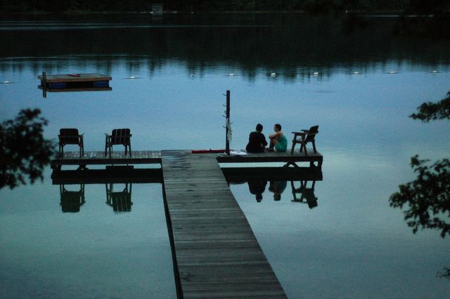A blue evening picture of two young women sitting on a dock over a ripple-dappled pond.