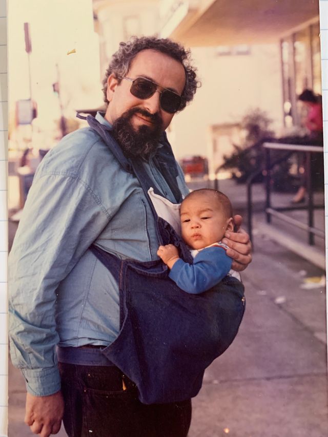 A vintage photograph of a man with dark curly hair, a full beard, and large dark sunglasses, wearing a denim jacket, cradling a baby in a blue outfit against his shoulder on a city sidewalk.