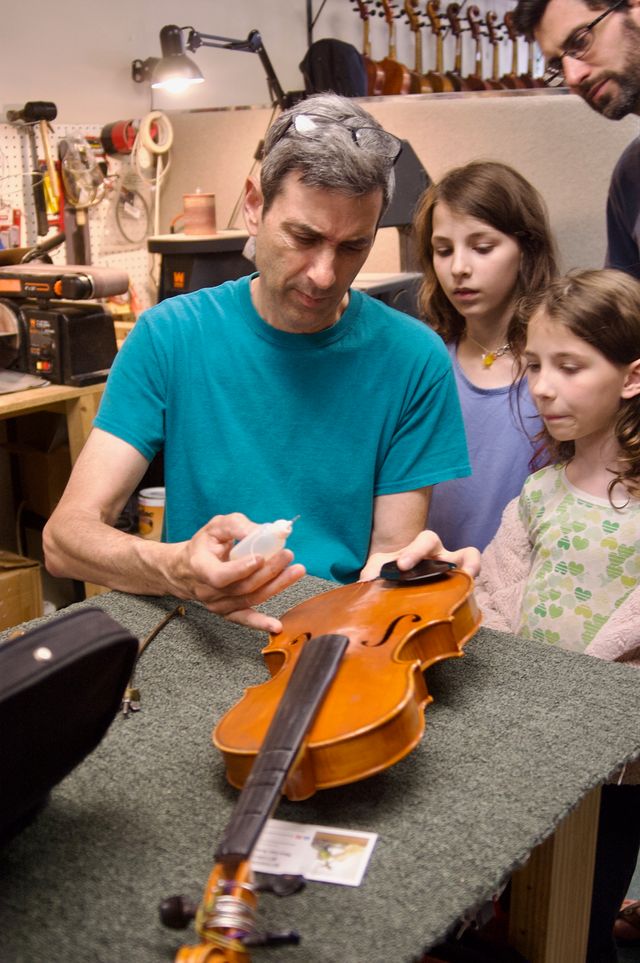 A man in a blue shirt holding an unstrung violin in one hand and a bottle of glue with a needlepoint in another hand while two young children and another man look on.