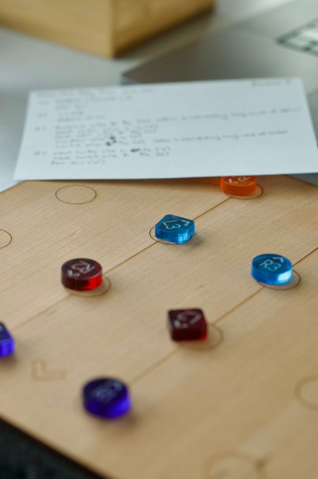 A close up a wooden board with dark circles engraved on it in a grid pattern. There are colorful plastic tokens on it labeled things like "L3," "R3," "L2," and "R2." The focus is very shallow. There's an index card with writing in the background but it's too blurry to make out.