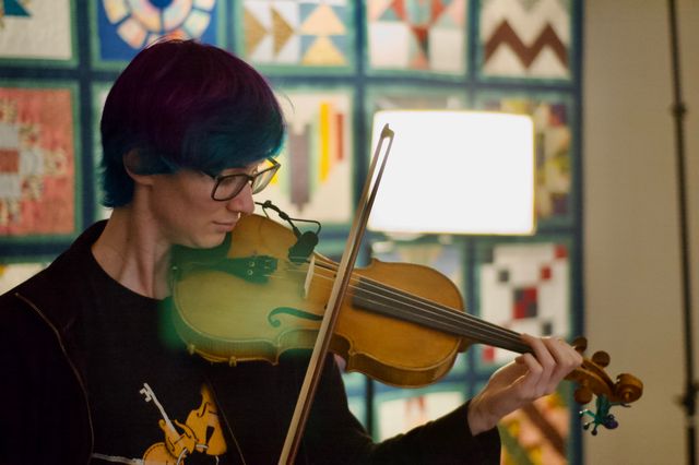 A woman with short colored hair, playing fiddle in front of a blurred quilt background. There's a lens flare in the foreground from a lamp visible behind her.