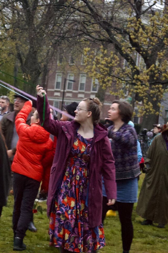 A crowd of people holding strands of colored ribbon attached to something high up off to the left of the frame. In the center of the frame a blonde woman in a colorful floral dress and purple jacket holds a green ribbon.