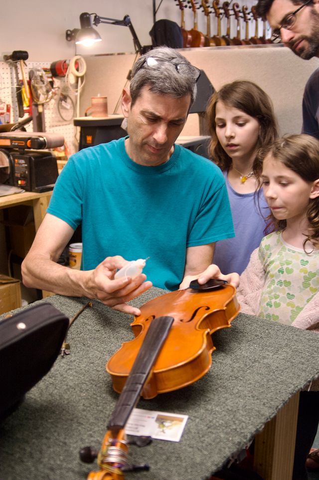 A man, intently working on a fiddle with no strings or bridge. In his other hand he's holding a bottle with a needlepoint cap. Behind him two young girls and their father look on with interest.