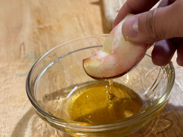 A close up of a hand holding a slice of red apple with pink tinged flesh. The slice is dripping from one end with honey over a small glass bowl full of honey.