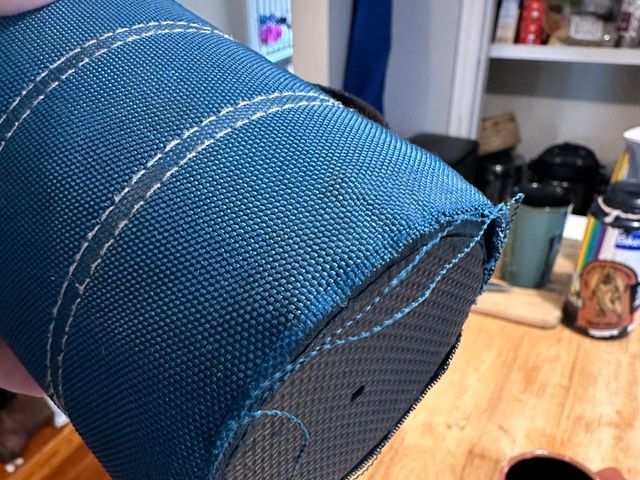A travel mug with a woven exterior. The outer fabric is blue and the base is detached from the plastic of the mug and there are fraying thread at the edges of the fabric.