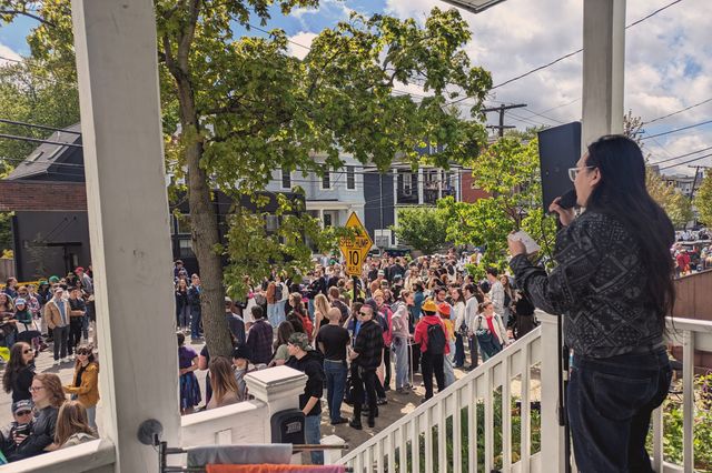 A street filled with people at some sort of festival as viewed from the porch of a house on that street. In the foreground, on the porch, a man with long black hair is standing at a microphone.