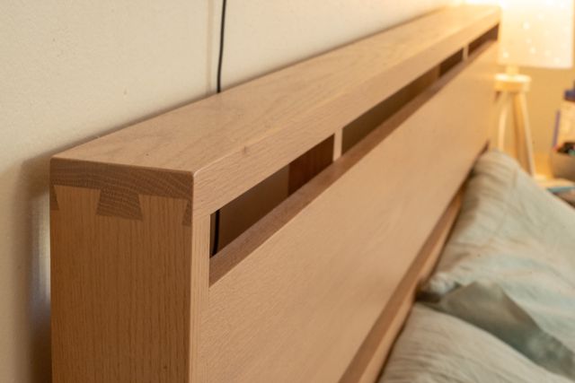 A close-up of the dovetail joint on the corner of the bed frame. Pillows and the far nightstand with a lamp can be seen in the background.