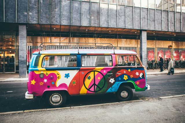 A VW bus on a city street painted in bright childlike rainbow colors with stars, flowers, and a large black peace sign in the center of the door.