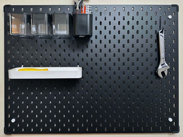 An IKEA Skådis board mounted on a wall. There are boxes and trays mounted on the board and an adjustable wrench hangs from a hook. On each corner of the board is a black bumper.