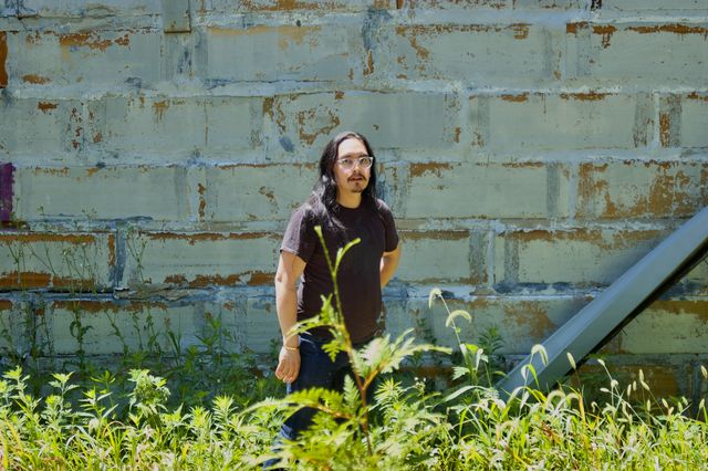 A 30-something Asian man with long hair against a distressed gray painted exterior wall. In the foreground, green brush is visible.