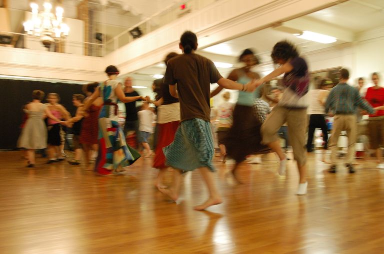 Dancers holding hands in a big circle, motion-blurred, in a hall with a polished wood floor and a chandelier. At the center a barefoot dancer with long dark hair tied back, wearing a brown t-shirt and a teal patterned skirt, has their back to the camera.