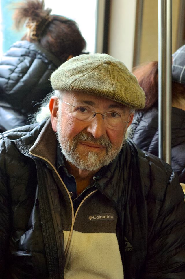 An older white man wearing a newsy cap, white and brown fleece, and black puffy jacket. He's looking directly at the camera with a neutral expression on his face. The background looks like a subway train.