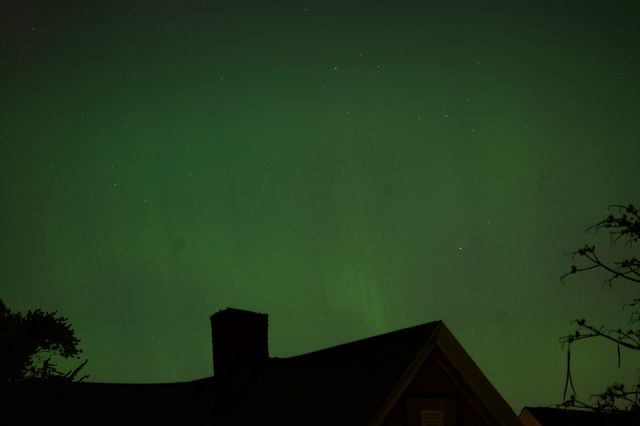 A green sky with white dotty stars hovers over the silhouette of the roof of a house and a couple trees. In the green sky some shimmering striations are vaguely visible.