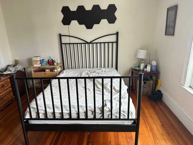 A bedroom, slightly cluttered, with a black metal bars bed frame in the middle. The bed is stripped down to sheets.