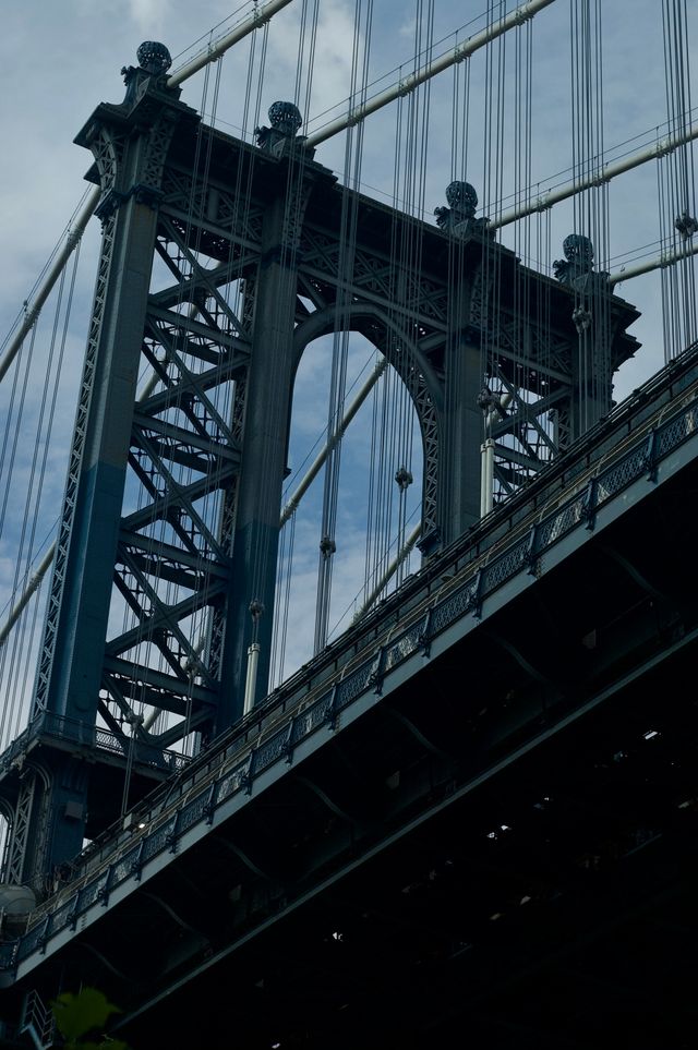 View from below of a dark green suspension bridge, cables and tower forming vertical lines crossed by the diagonal line of the deck seen from below.