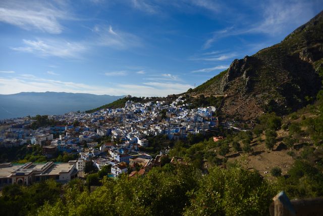 Distant view of a Moroccan town nestled into a green mountainside on a sunny day. The town’s buildings are painted varying shades of blue or beige with red roofs.