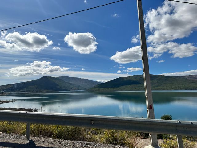 A body of water with green hills behind it, as seen from a freeway. The sky is blue with fluffy cumulus clouds.