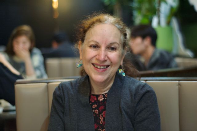 An older white woman with folded paper crane earings, a dark gray cardigan, and a red floral shirt. She's looking off to the right and smiling.